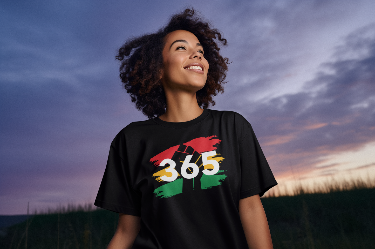 A woman with curly hair wears a black "365 Tee" from Sharp Tact Kreativ, made of soft ring-spun cotton featuring a pan-African design. She stands outdoors under the cloudy sunset sky, exuding style and comfort.