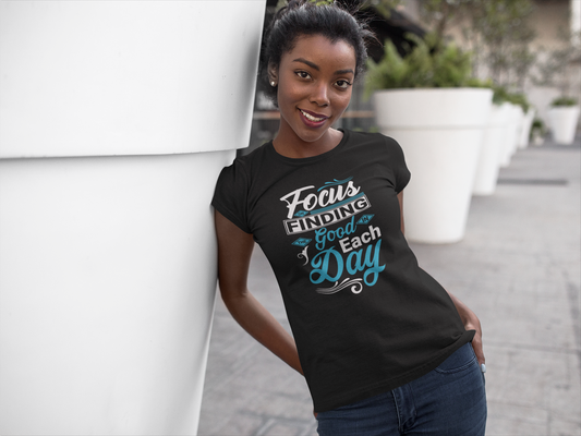 A woman smiles, leaning against a large planter, wearing Sharp Tact Kreativ's Focus on Finding the Good in Each Day Tee that encourages positive living.