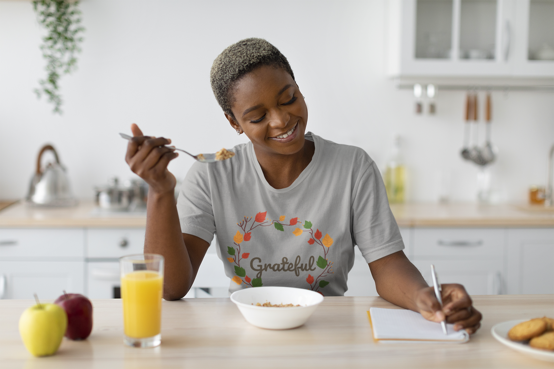 A woman in a comfortable Grateful Tee sweatshirt, made by Sharp Tact Kreativ, is writing on a notebook while eating breakfast, reminding herself of important information.