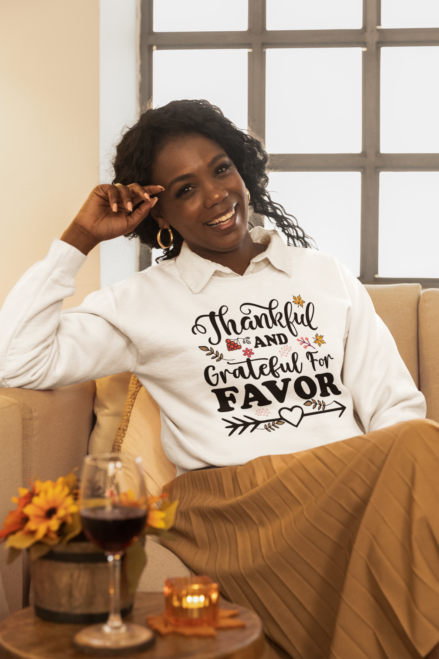 A woman sitting on a couch wearing the Thankful and Grateful for Favor Sweatshirt by Sharp Tact Kreativ, a cotton blend sweatshirt in crewneck style.