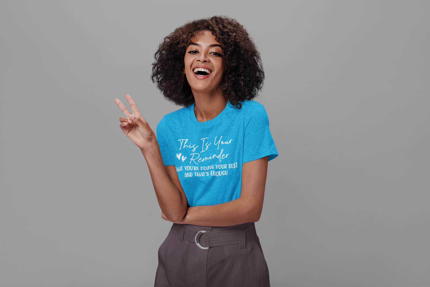 A woman wearing the "This is Your Reminder that You're Doing Your Best and That's Enough Tee (heart)" by Sharp Tact Kreativ | Tees & Gifts with Encouraging Messages to Brighten Your Day with a Bit of Wit posing for the camera.