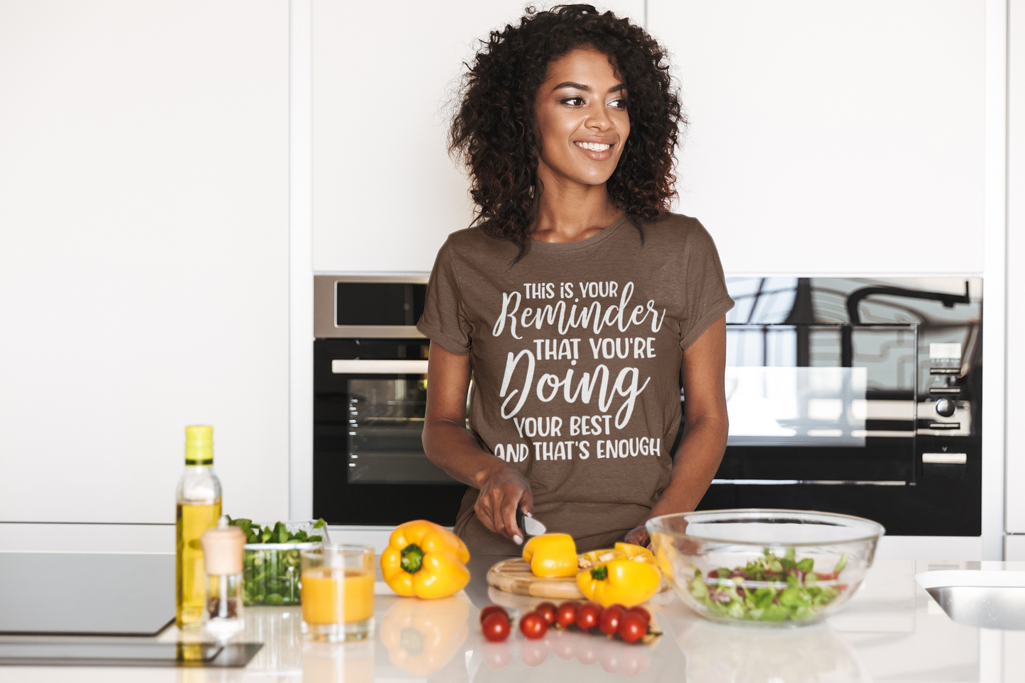 A woman in a kitchen preparing food while wearing the "This is Your Reminder that You're Doing Your Best and That's Enough Tee" from Sharp Tact Kreativ | Tees & Gifts with Encouraging Messages to Brighten Your Day with a Bit of Wit.
