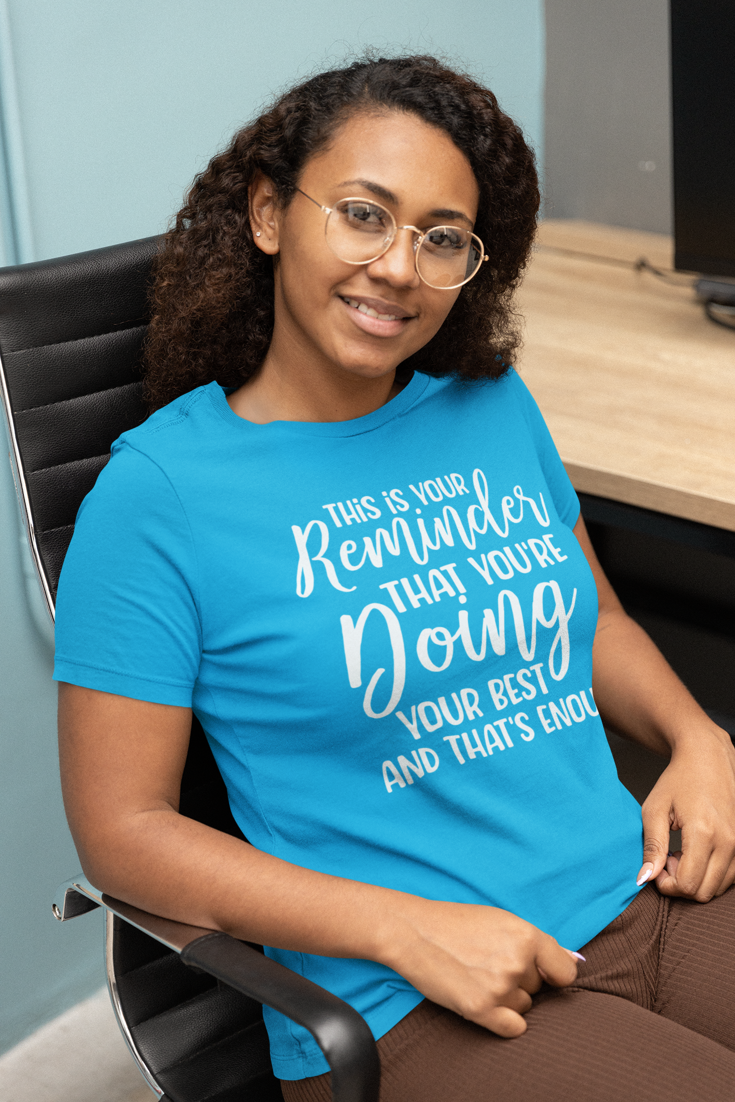 A woman sitting in a chair wearing a Sharp Tact Kreativ | Tees & Gifts with Encouraging Messages to Brighten Your Day with a Bit of Wit t-shirt that says "This is Your Reminder that You're Doing Your Best and That's Enough Tee".