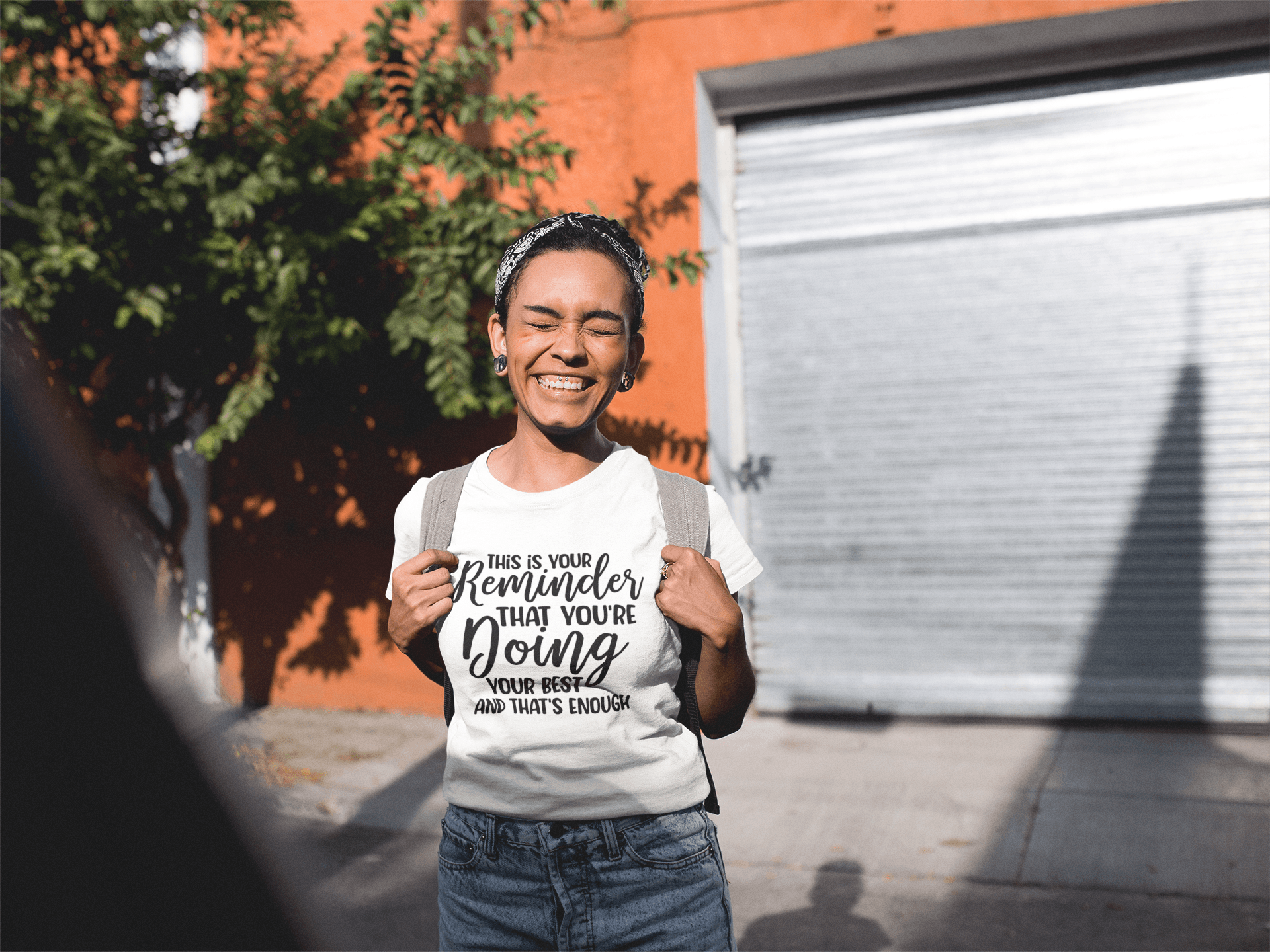 A young woman wearing a white "This is Your Reminder that You're Doing Your Best and That's Enough Tee" t-shirt and a backpack.