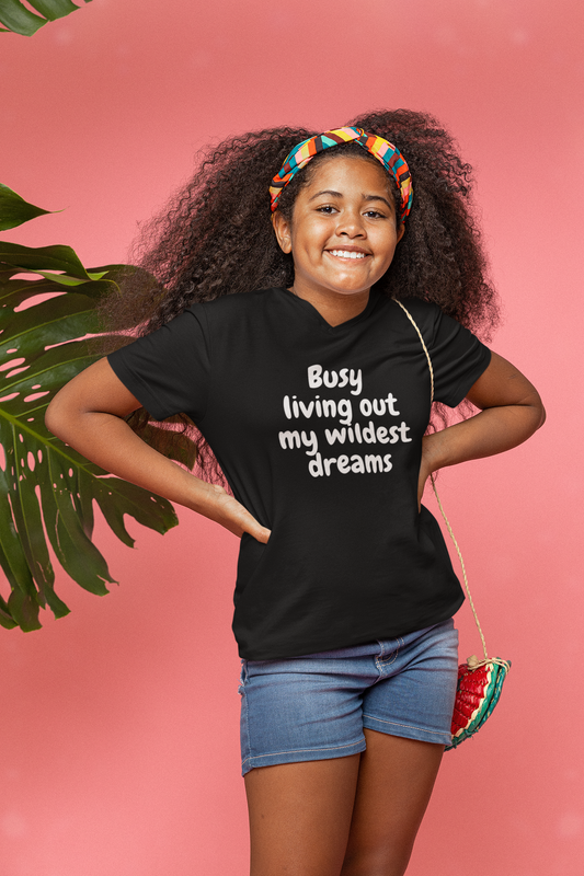 A young girl wearing a black t-shirt that says, Busy Living Out My Wildest Dreams Tee (Youth Size) by Sharp Tact Kreativ | Tees & Gifts with Encouraging Messages to Brighten Your Day with a Bit of Wit.