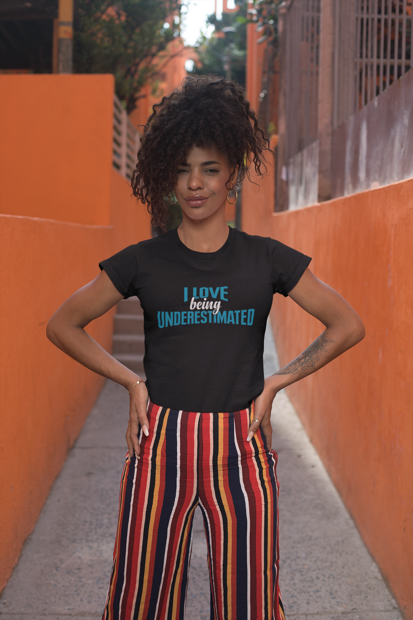A woman wearing a black "I Love Being Underestimated" t-shirt by Sharp Tact Kreativ and striped pants.